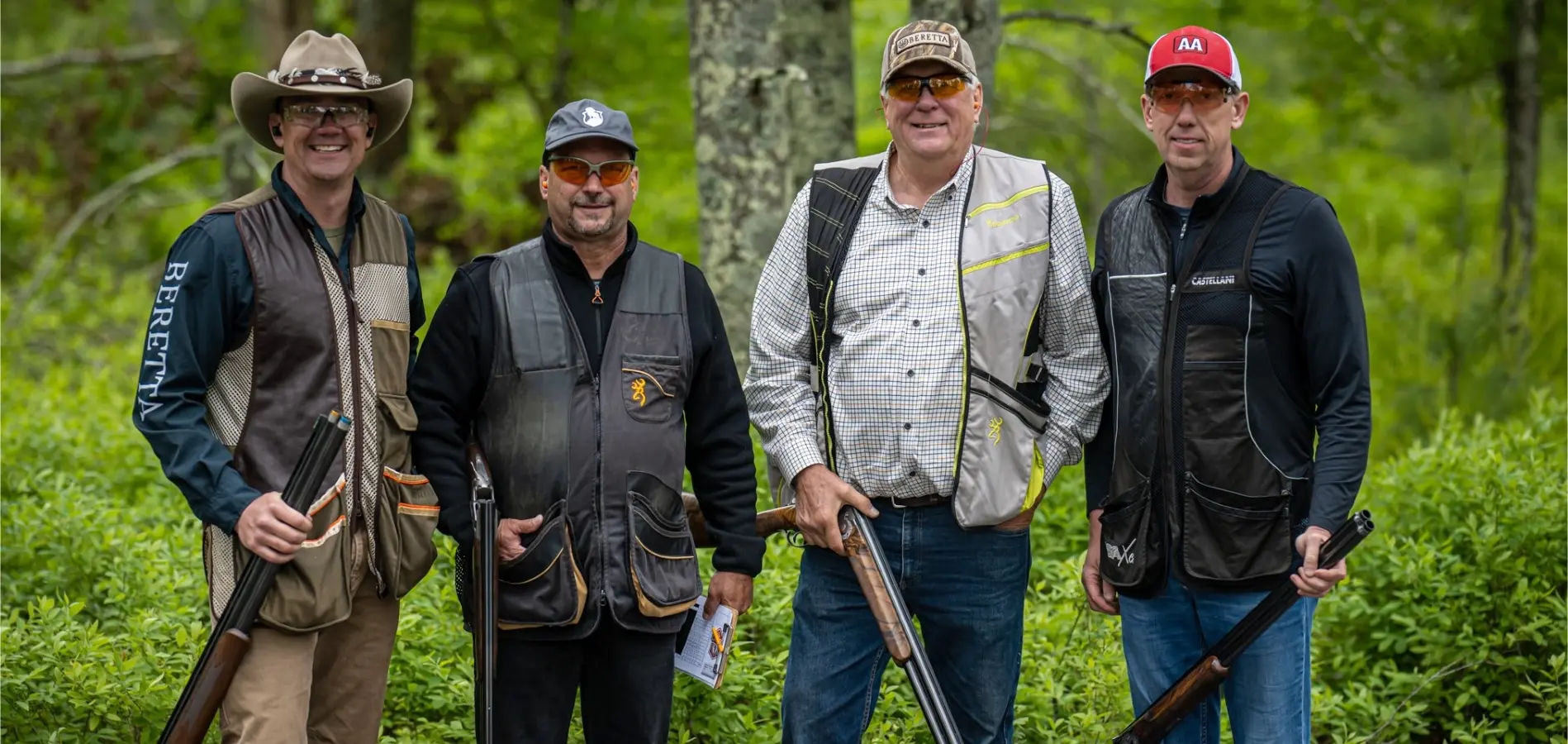 Four men in shooting vests and gear posing with shotguns in a scenic, wooded area at The Preserve Sporting Club, highlighting outdoor activities